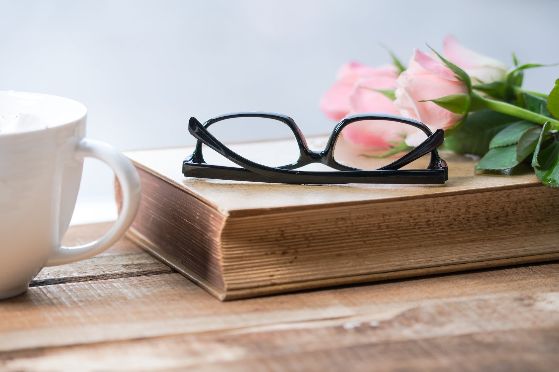 eyeglasses on book beside pink rose on cup