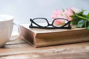 eyeglasses on book beside pink rose on cup